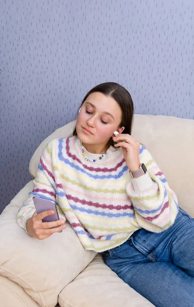 Jeune fille à la mode rêveuse se détendre sur un canapé, en utilisant smartphone regarder la vidéo, mettre des écouteurs. — Photo