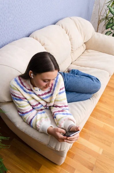 Álmodozó tinédzser lány pihen a kanapén, okostelefont használ videónézéshez fülhallgatóval. Zenehallgatás online. — Stock Fotó