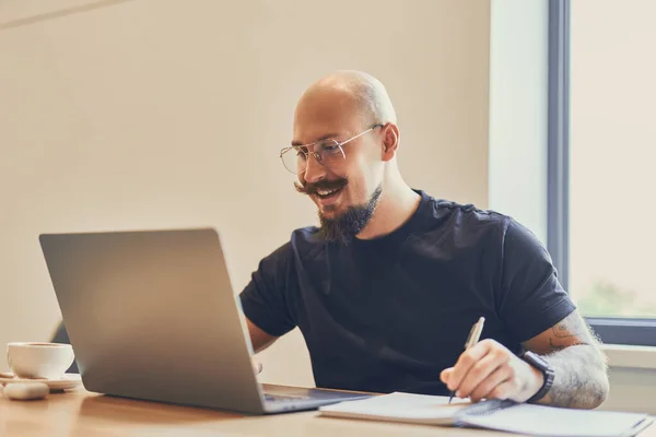 Молодий кавказький фрілансер працює з ноутбуком за своїм столом в офісі, роблячи щось нотатки — стокове фото