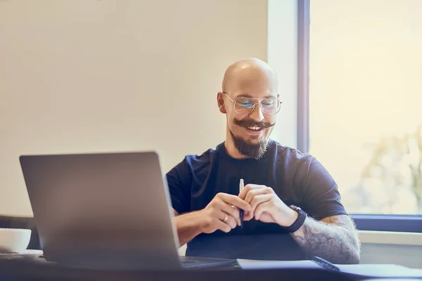 Smiling caucasian bald man manager using laptop while writing notes in notebook at workplace.