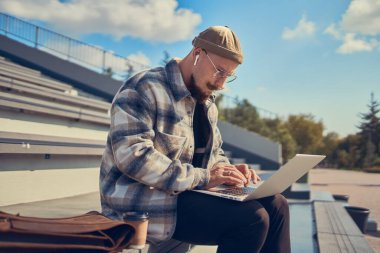 Konsantre sakallı adam dışarıda oturuyor ve dizüstü bilgisayarında yazı yazıyor. 4G internet bağlantısı