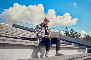 Genç adam dizüstü bilgisayar kullanıyor, serbest çalışan dışarıda çalışıyor, klavyede daktilo çalıyor, bilgisayarla sohbet ediyor..