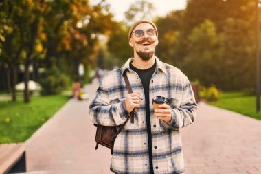 Yakışıklı, mutlu, sakallı adam şehir parkında yürürken dışarıda kahve içiyor. 