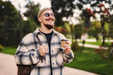 Şapkalı ve güneş gözlüklü olumlu adam sırt çantasıyla şehir merkezinde yürürken kahve tutuyor. 