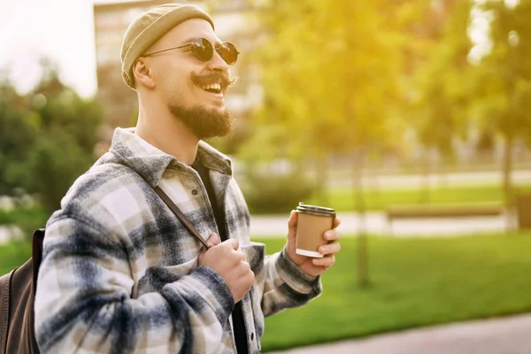 Handsome happy smiling bearded man drinks coffee outside the street while walks in the city park