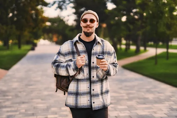 ハンサムな髭のスタイリッシュなヒップスターは、リラックスした時間を過ごしながら行くためにコーヒーと公園の屋外を歩きます — ストック写真