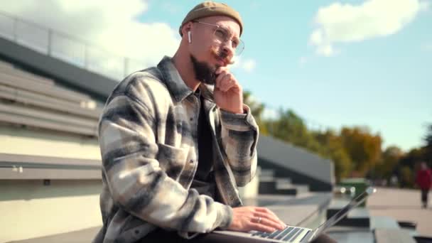 Schöner bärtiger Student mit Brille studiert, während er in der Nähe der Universität am Laptop sitzt — Stockvideo