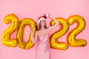 Gülümseyen yetişkin kız elinde Noel Baba şapkasıyla şampanya tutuyor. 2022 altın hava balonları yeni yıl konsepti. 