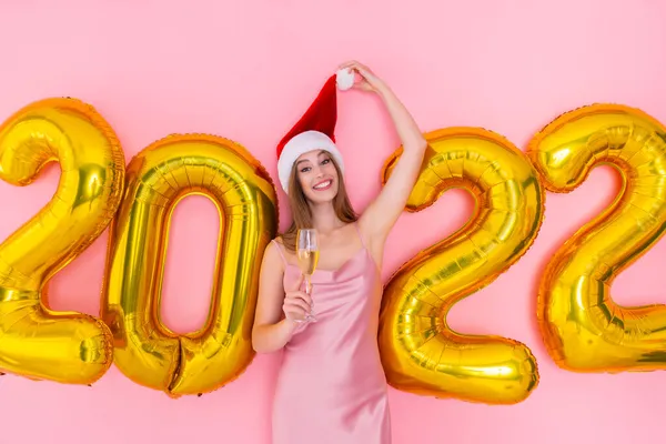 Rapariga sorridente levanta uma taça de champanhe no chapéu do Pai Natal. balões de ar dourado 2022. Festa de Ano Novo — Fotografia de Stock