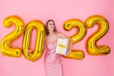 Mutlu kız 2022 hava balonlarının yanında duruyor. Yeni yıl kutlaması için hediye kutusunu pembe arka planda tutuyor.
