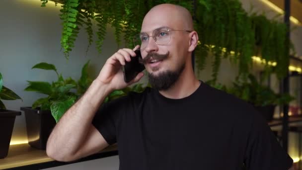 Atractivo joven barbudo hombre en vestidor casual hablando en el teléfono inteligente en el restaurante, conexión móvil — Vídeos de Stock