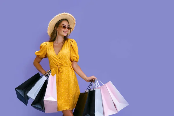 Boldog nő napszemüvegben és kalapban színes bevásárlószatyrokat tart lila háttéren. Értékesítési koncepció — Stock Fotó