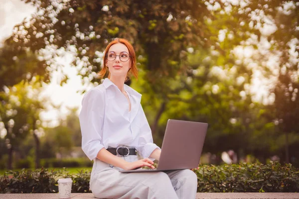 Студент рудої дівчини в окулярах і біла сорочка сидить в парку, використовуючи ноутбук з кавою, щоб піти — стокове фото