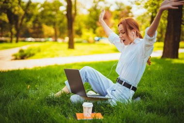 Kızıl saçlı duygusal kızın portresi parkta otururken dizüstü bilgisayarı açık havada eğleniyor.