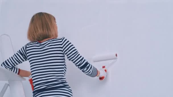 Πίσω όψη της γυναίκας ζωγραφίζει τον τοίχο σε λευκό χρώμα, ενώ στέκεται στις σκάλες. Σχέδιο ανακαίνισης — Αρχείο Βίντεο