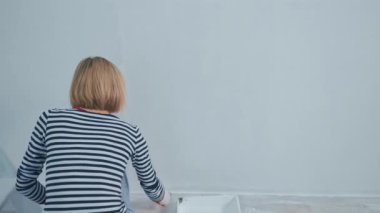 Sarışın kadının arka görünüşü, duvarı beyaz boyalı bir fırçayla boyuyor. yenileme konsepti