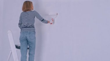 Kadının arka görüntüsü merdivenlerde dururken fırça rulosuyla duvarı boyuyor. Ev Yenileme