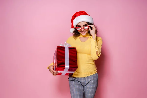 Sonriente chica rizada en sombrero de Santas, gafas sostiene presente mientras está de pie sobre fondo rosa. Año Nuevo — Foto de Stock
