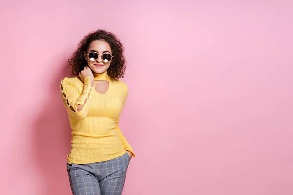 Menina encaracolado bonita sorrindo em roupa amarela e óculos de sol enquanto posando no fundo do estúdio rosa — Fotografia de Stock