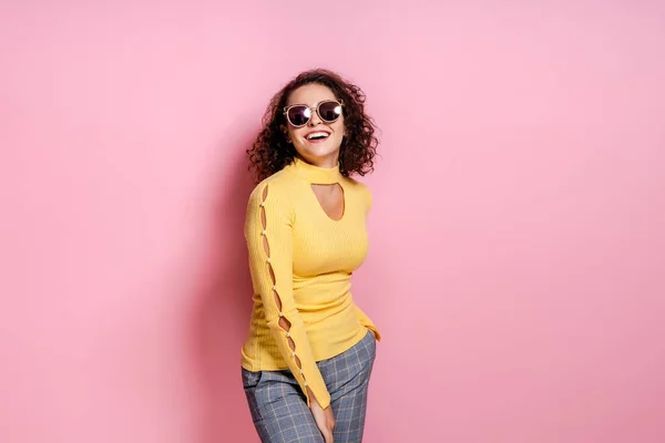 Rindo menina encaracolado vestido roupa amarela e óculos de sol enquanto posando isolado sobre fundo rosa. — Fotografia de Stock