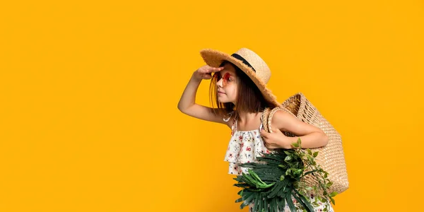 Симпатична маленька дівчинка в солом'яному капелюсі в сонцезахисних окулярах тримає сумку з тропічними рослинами щось виглядає — стокове фото