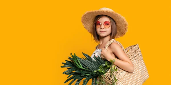 Симпатична маленька дівчинка в солом'яному капелюсі, помаранчеві сонцезахисні окуляри тримають солом'яну сумку з тропічними рослинами . — стокове фото