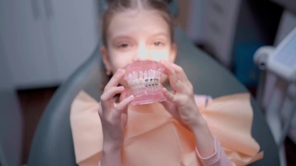 Красивая маленькая девочка улыбается, держит модель челюсти в то время как сидит в стоматологическом кресле в клинике весело — стоковое видео