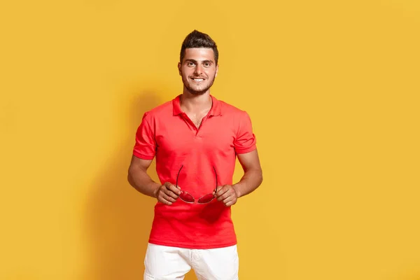 Joven hombre hipster en camiseta roja y pantalones cortos blancos sostiene gafas de sol mientras está parado sobre fondo amarillo — Foto de Stock