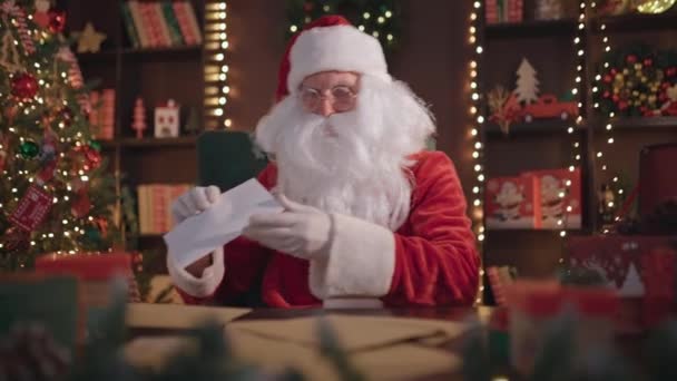 Счастливый Санта-Клаус читает письма, смотрит и разговаривает на камеру, поздравляет сидя за столом дома — стоковое видео