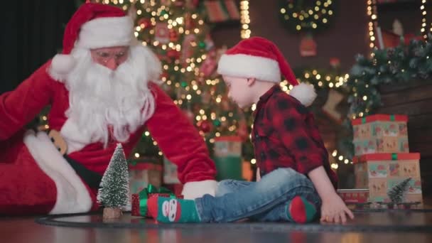 Vidéo de Petit garçon en bas âge jouant avec le Père Noël sur le sol sous l'arbre de Noël avec locomotive — Video
