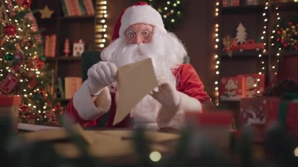 Эмоциональный шокированный Санта Клаус проверяет конверты с рождественскими пожеланиями, разговаривая с камерой — стоковое видео