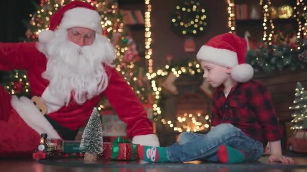 Відео маленького хлопчика, який грає з Санта Клаусом на підлозі під ялинкою з залізницею. — стокове відео
