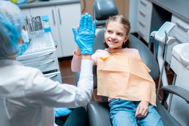 Dost canlısı dişi dişçi, tedavi sonrası Stomatolojik sandalyede oturan küçük kıza beşlik çakıyor.