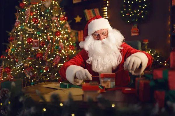 Der Weihnachtsmann oder der Nikolaus macht Geschenke, während er zu Hause am Tisch neben dem Weihnachtsbaum sitzt — Stockfoto