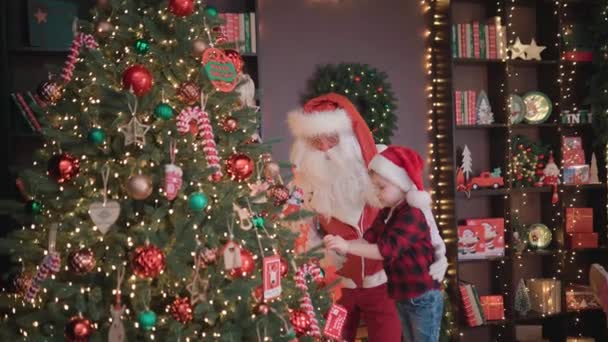 Święty Mikołaj z małym dzieckiem udekorować choinkę w przytulnym domu w pokoju z uroczystym wnętrzem. — Wideo stockowe