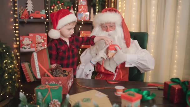 Święty Mikołaj i mały asystent chłopca robi prezenty świąteczne dla dzieci. Boże Narodzenie NOWY ROK DOTYCZĄCY — Wideo stockowe