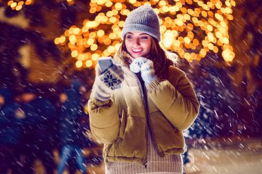 Gülümseyen mutlu kız dışarıda Noel süslerinin önünde telefonu duygusal olarak kullanıyor.