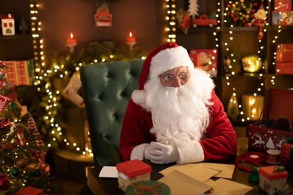 Der Weihnachtsmann sitzt zu Hause am Tisch und macht sich bereit, Briefe mit Weihnachtswünschen zu lesen — Stockfoto