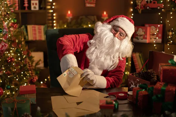 Der Weihnachtsmann bereitet zu Hause am Schreibtisch Geschenke für Kinder für Weihnachten vor, während er einen Brief zückt — Stockfoto