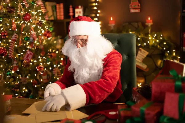 Der Weihnachtsmann bereitet zu Hause am Schreibtisch Geschenke für Kinder für Weihnachten vor, während er Wunschzettel öffnet — Stockfoto