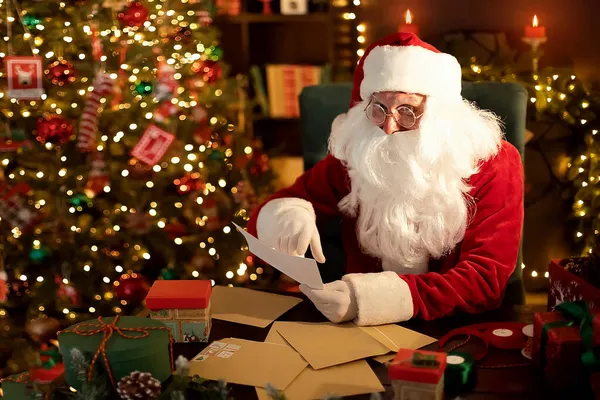 Der Weihnachtsmann bereitet am Schreibtisch Geschenke für Kinder für Weihnachten vor, während er zu Hause Wunschzettel liest — Stockfoto