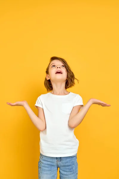 Емоційна маленька дівчинка в білій футболці дивиться вгору, жестикулюючи руками на жовтому тлі — стокове фото