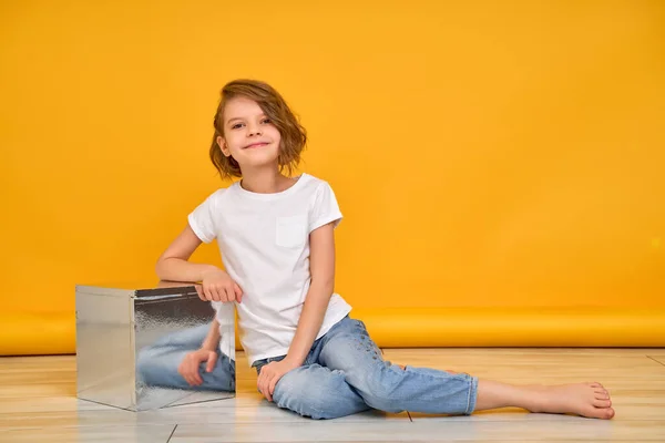 흰색 티셔츠와 청바지를 입은 어린 소녀가 노란 배경 위에 포즈를 취하는 동안 바닥에 앉아 있다 — 스톡 사진