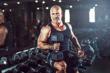 Kel erkek vücut geliştirici karanlıkta spor salonunda poz verirken kollarında halterler tutuyor. Vücut geliştirme kavramı.