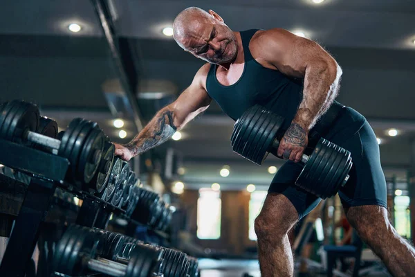 Spor salonunda poz verirken dambıllarla egzersiz yapan yaşlı bir vücut geliştirici. Vücut geliştirme kavramı.