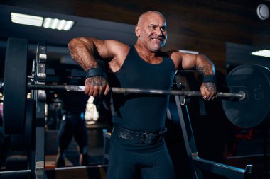 Duygusal yaşlı kel vücut geliştirici spor salonunda halterle trapezius ve deltoid egzersizi yapıyor.