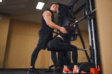 Genç kadın, eğitmenle spor salonunda halterle bacak egzersizi yapıyor. Çömelme egzersizi. Kişisel antrenör