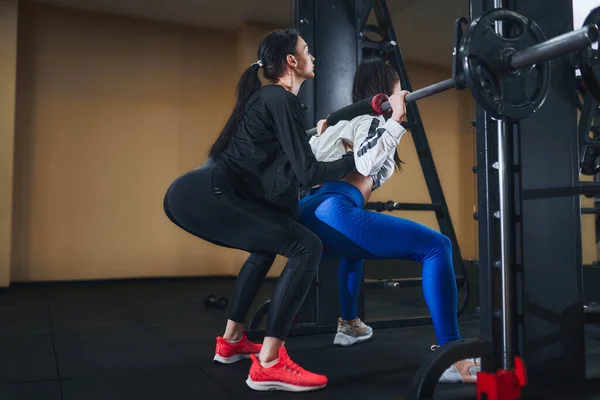 Jonge brunette die benen traint met lange halter in de sportschool. persoonlijke vrouwelijke trainer. Krachtoefening — Stockfoto