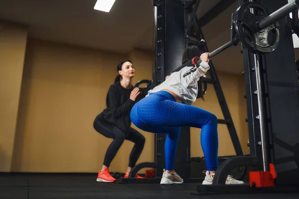Jonge blanke vrouw die benen traint met lange halter in de sportschool met instructeur. Persoonlijke vrouwelijke trainer — Stockfoto