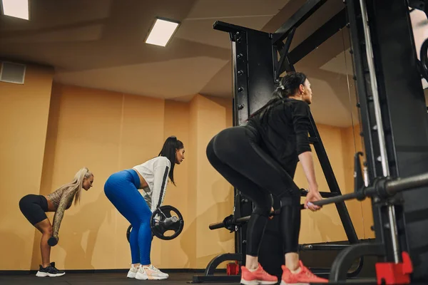 Sportieve meisjes met rugleuning naar de camera uitvoeren van deadlift training activiteit met lange halter in de sportschool — Stockfoto